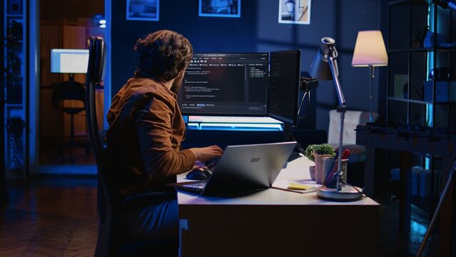 Software programmer putting headphones on while developing code, making time pass faster during remote job shift. IT professional doing multitasking in apartment office while listening music, camera B