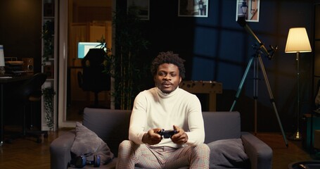 Gamer arriving home, ready to start game and enjoy leisure time. African american man sitting on couch in living room, playing videogame on gaming console, relaxing after work