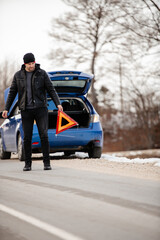 Man putting triangle caution sign on road near broken car.