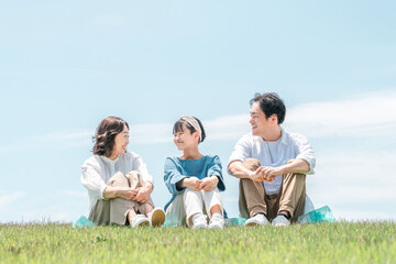 青空の見える公園・丘に座る家族・親子（父親・母親・娘）
