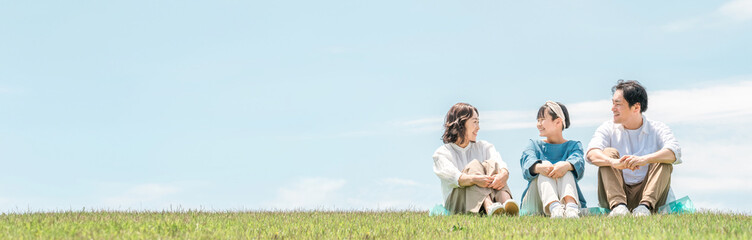 Fototapeta premium 青空の見える公園・丘に座る家族・親子（父親・母親・娘） 