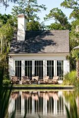 Obraz premium A small white cottage with black windows and doors, a shingle roof, and tall trees behind it, surrounded by lush landscaping and sitting atop an empty lagoon. Large wooden outdoor furniture.