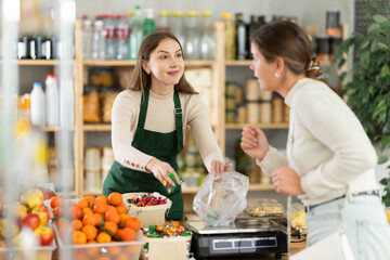 Grocery store selling area. Female employee sells candies to woman, puts them in bag, puts them on scale and weighs sweets. Brunette European girl buys candies by weight.