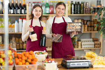 Young women sellers stands in trading hall of store with goods in hand, wearied in apron stands in trading hall of minimarket, ready to greets visitors and offer wine with sweet snacks in selling area