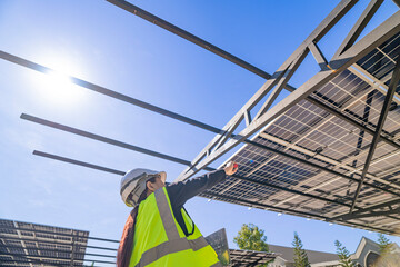 A technician in a safety helmet and reflective vest inspects a solar panel structure using a...