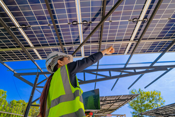 Fototapeta premium A technician in a safety helmet and reflective vest inspects a solar panel structure using a laptop. The solar panels are mounted on a steel carport framework under a clear blue sky.