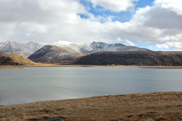 A huge lake with gently sloping shores and yellowed grass at the foot of a high mountain range with snow-capped peaks.
