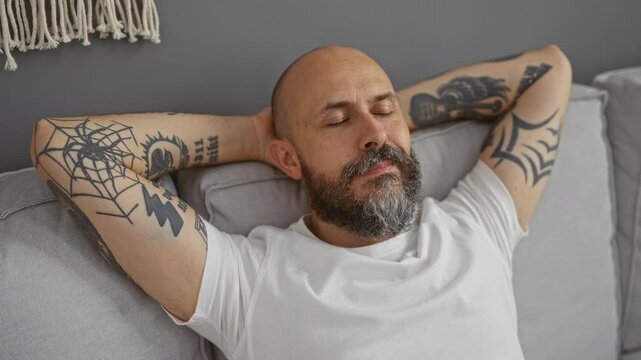Handsome man with beard and tattoos relaxing indoors with closed eyes wearing white shirt in modern living room