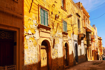 Old weathered street in Malta with golden buildings, peeling paint, arched wooden doors, and green shuttered windows. Shadows cast by sunlight add depth to the rustic Mediterranean atmosphere