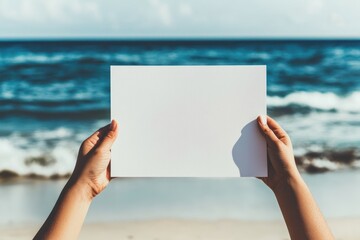 A person holding a blank white canvas by the beach, with waves and ocean in the background.