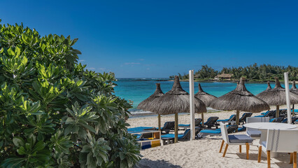 Rows of deck chairs with mattresses under straw umbrellas on the sandy beach. There are wicker chairs, a table on the embankment. Calm turquoise ocean, blue sky. A tropical shrub in the foreground. 