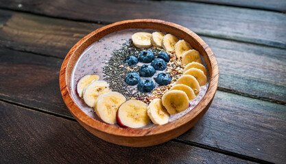 Fototapeta premium A wooden bowl filled with a creamy vegan smoothie, elegantly topped with fresh banana slices, ripe blueberries, and chia seeds, resting on a rustic wooden table