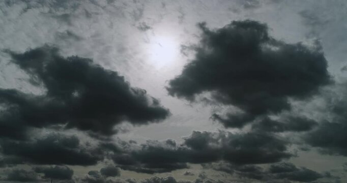 Timelapse of the overcast sky with the sun softly shining through light grey altostratus clouds and contrasting dark cumulus clouds moving fast and changing shapes