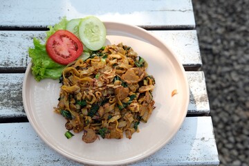 A delicious plate of fried flat rice noodles (kwetiau goreng) served with fresh cucumber, tomato, and lettuce garnish, stir-fried with vegetables, soy sauce, and spices for an authentic Asian flavor