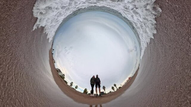 A "tiny planet" perspective captures a man and a woman standing on a sandy beach as waves encircle the spherical view. The immersive effect emphasizes the motion of the approaching water and the open 