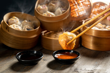 Hand with chopstick eating Chinese dim sum dumplings on bamboo wooden basket bowl with sauce and...