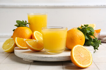 Wooden board with glasses of tasty juice, oranges and mint leaves on white tile table