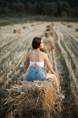 the girl looks to the side, and her hands lie on her knees. More haystacks and a blurry field landscape are visible in the background.