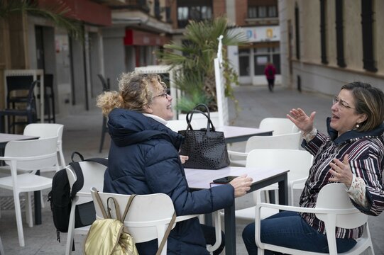 Two middle-aged friends enjoying a cup of coffee on a terrace in the main square of a Spanish village, chatting and laughing.