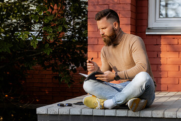 Stylish bearded man in brown turtleneck sweater and jeans sitting in lotus pose on beton steps...