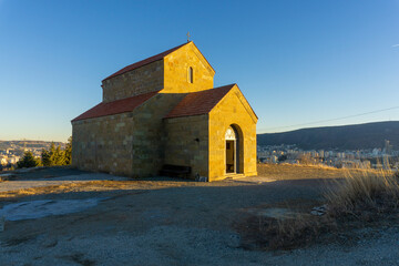 Fototapeta premium A small stone church on a hill above the city of Tbilisi is illuminated by the warm evening sunlight. Part of the city and mountains are in the background.