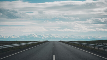 Vast Asphalt Road Stretching into the Horizon with Clear White Dividing Lines and Metal Guardrails, Surrounded by Majestic Snow-Capped Mountains, Fluffy Clouds in the Sky, Evoking Adventure and Explor