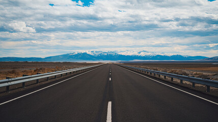 Vast Asphalt Road Stretching into the Horizon with Clear White Dividing Lines and Metal Guardrails, Surrounded by Majestic Snow-Capped Mountains, Fluffy Clouds in the Sky, Evoking Adventure and Explor
