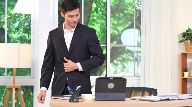Handsome businessman looking out window then walking towards desk and continuing to work on laptop modern office