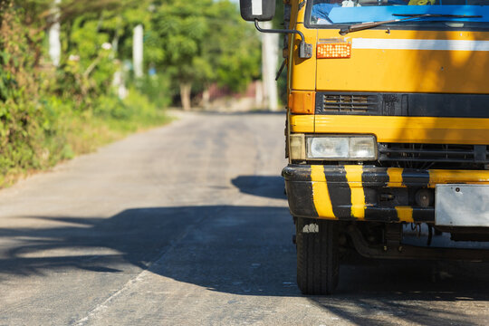 Front of a yellow truck parked on a paved road. The truck features black-and-yellow hazard stripes on the bumper, a large rectangular headlight, and an indicator light