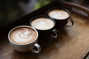 Three cups of beautifully crafted latte art placed on a wooden tray, each showcasing intricate milk foam designs, creating a cozy and inviting atmosphere perfect for a coffee break or gathering