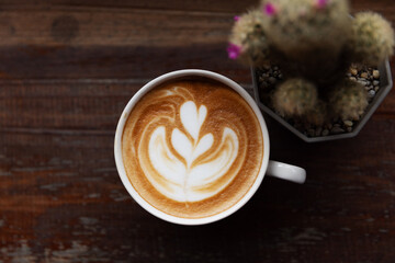 A beautifully crafted cup of latte with intricate heart-shaped foam art sits on a rustic wooden table, accompanied by a small potted cactus, creating a cozy and artistic coffee moment