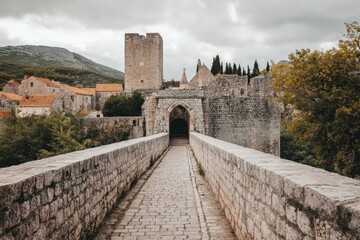 Obraz premium A historic stone bridge extends towards a medieval fortress under a cloudy sky, surrounded by lush greenery and mountainous landscape, evoking timelessness and mystery.