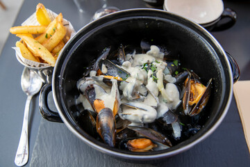 Gros plan sur des moules-frites sauce roquefort aux herbes, servies avec des frites croustillantes sur une table sombre avec une grosse cuillère. En terrasse d'un restaurant français.