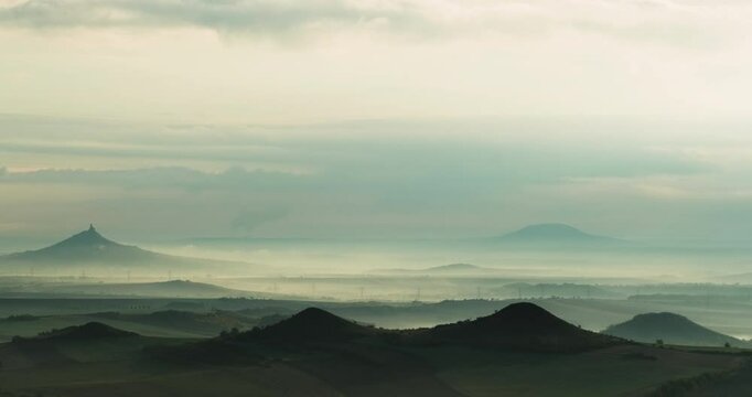 Timelapse of hilly landscape with a silhouette of castle ruins on one hillwith mist rolling in valleys and shadows transforming with time and of overcast sky white and grey clouds on a cloudy day 