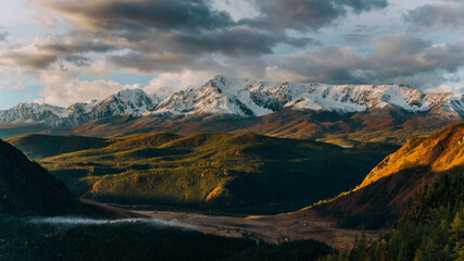 Golden hour sunlight illuminates a breathtaking panorama of snow-capped mountains towering over rolling green hills and a tranquil valley, creating a stunning display of nature's beauty