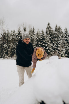 A happy young father plays with his little son and throws him into the snow. Both are dressed in warm winter clothes. The background is a large snowy forest.