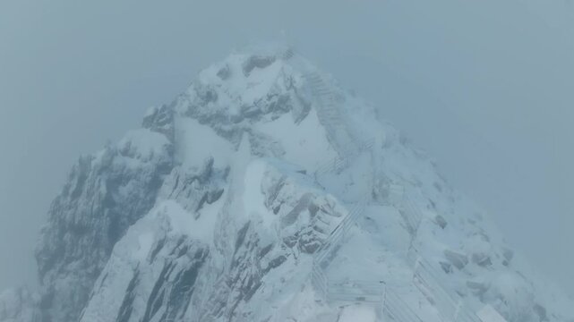 Aerial view of a snow-covered mountain peak - Lomnicky stit (Slovakia) surrounded by dense fog, showcasing dramatic rock formations and icy winter landscapes. (4K)