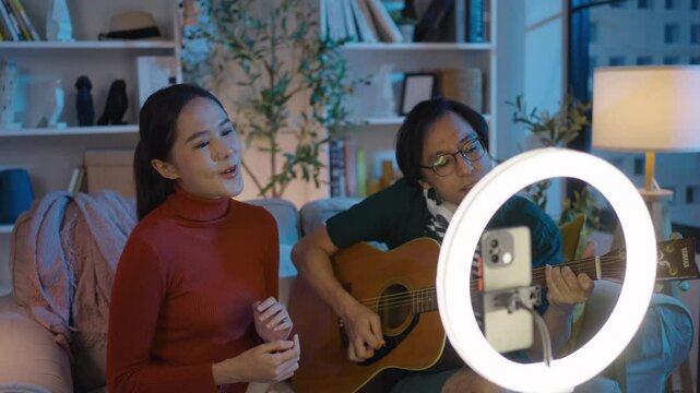 Asian couple live-streaming a singing session while playing guitar on social media in their living room at night.