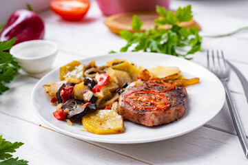 Toasted beef steaks with vegetables on a white plate. Grilled pork steaks with potatoes and vegetables on a white plate on a white table. A hearty lunch. Close-up