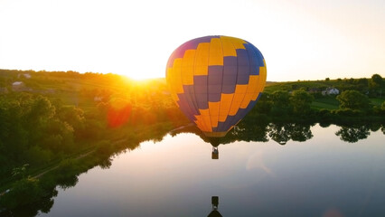 Hot air balloon floating over lake at sunrise. A vibrant hot air balloon floats over a calm lake at sunrise, with the golden light reflecting on the water.