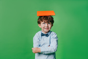 Happy preschool boy wearing bow tie holding book on the head standing over green background