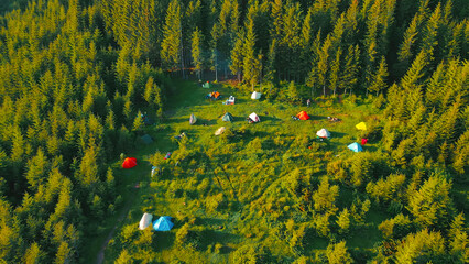 Campsite with colorful tents in a forest clearing. An aerial view of a campsite set in a forest clearing, with various colorful tents scattered across the lush green grass. © Iryna
