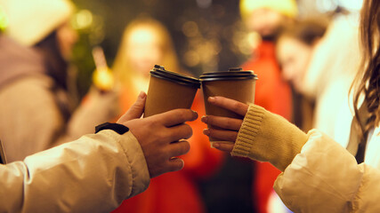 Close-up of two disposable coffee cups being toasted together at cozy winter gathering with blurred festive lights in the background. Concept of winter holidays, Christmas, traditions