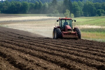 Fototapeta premium A red tractor is plowing a large brown field, preparing it for planting. The scene is set against a backdrop of lush green trees and a cloud-dotted sky.