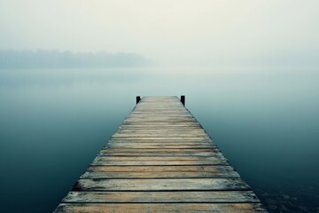 Fototapeta premium A tranquil wooden pier extends into a serene, mist-shrouded lake, evoking feelings of peace and solitude, framed by an ethereal horizon in this reflective moment.