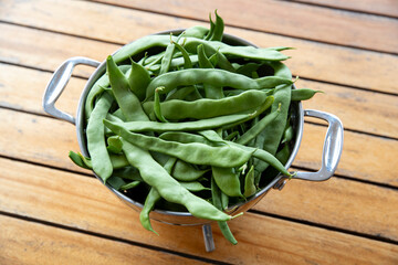 Récolte de haricot mangetout frais dans une passoire métallique posée sur une table en bois.