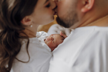 A couple is kissing a baby in a white shirt. The baby is wrapped in a blanket and is being held by the man. The woman is kissing the baby's face