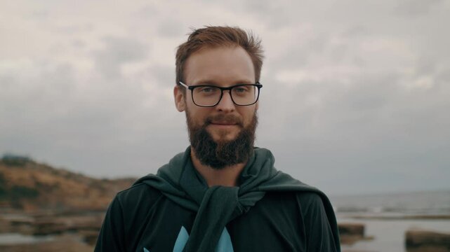 Portrait smiling bearded man in eyeglasses posing at sea beach landscape with dramatic sky slowmo