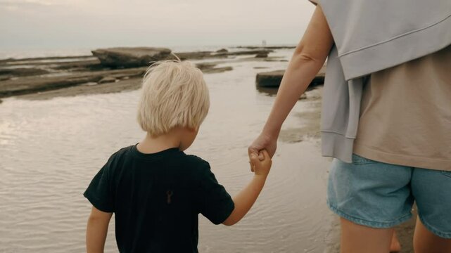 Cute male kid holding mother hand walking together at sea beach coastline travel vacation closeup