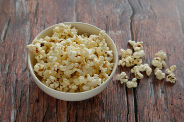 A white bowl filled with freshly popped popcorn placed on a rustic wooden table. A few scattered popcorn pieces add a casual and inviting touch to the scene.